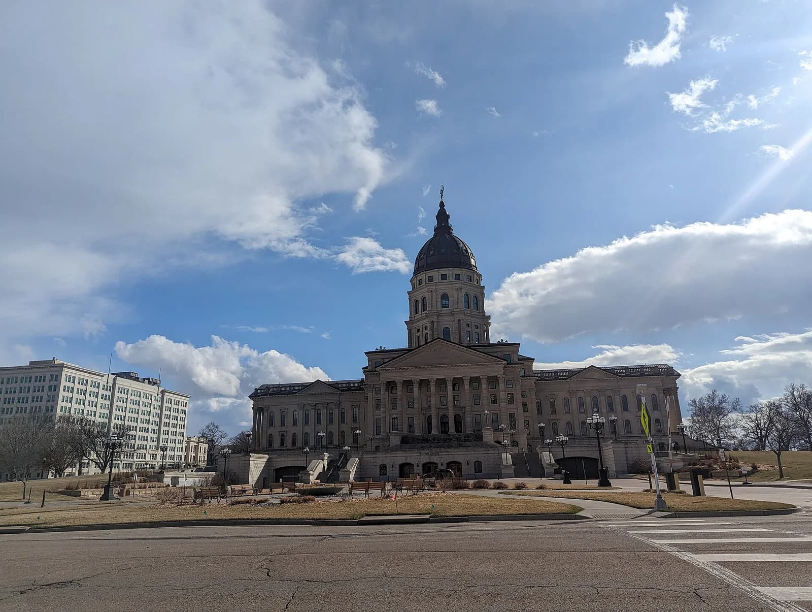 A large, classical-style building with a prominent dome stands under a partly cloudy sky. A modern, multi-story building is visible in the background to the left, and a street with a crosswalk is in the foreground.