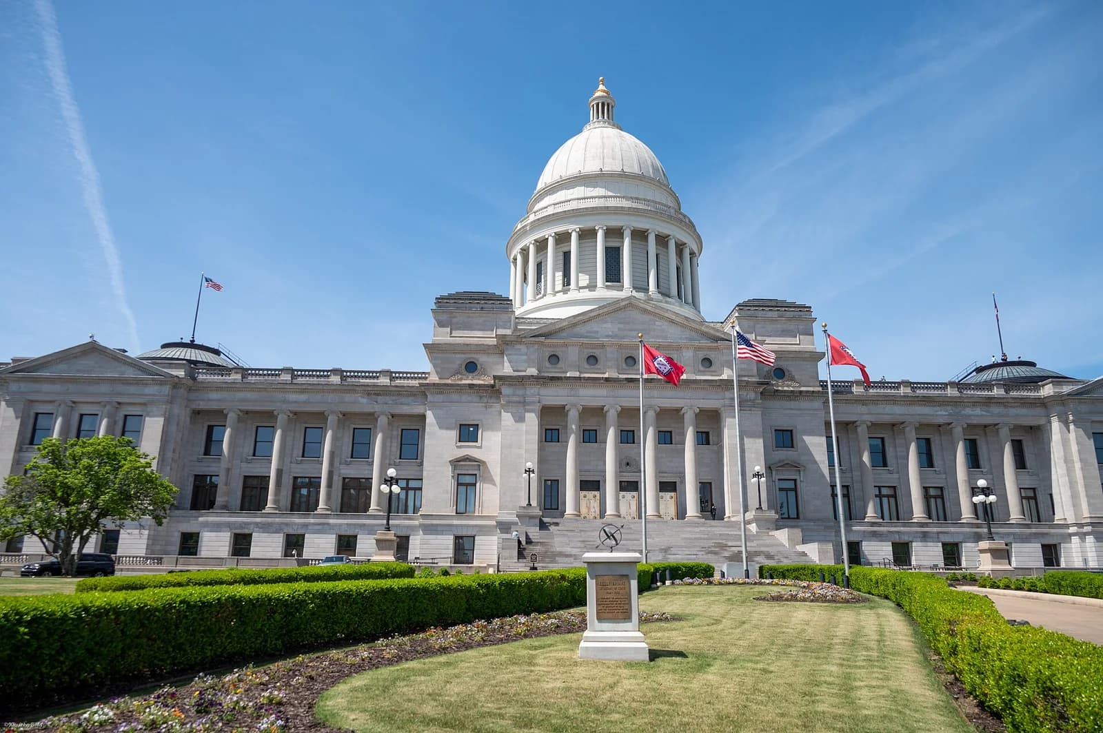 A grand, light-colored stone building with a large central dome and columns stands under a clear blue sky. Manicured green lawns and hedges are in the foreground, with flags flying near the building's entrance.