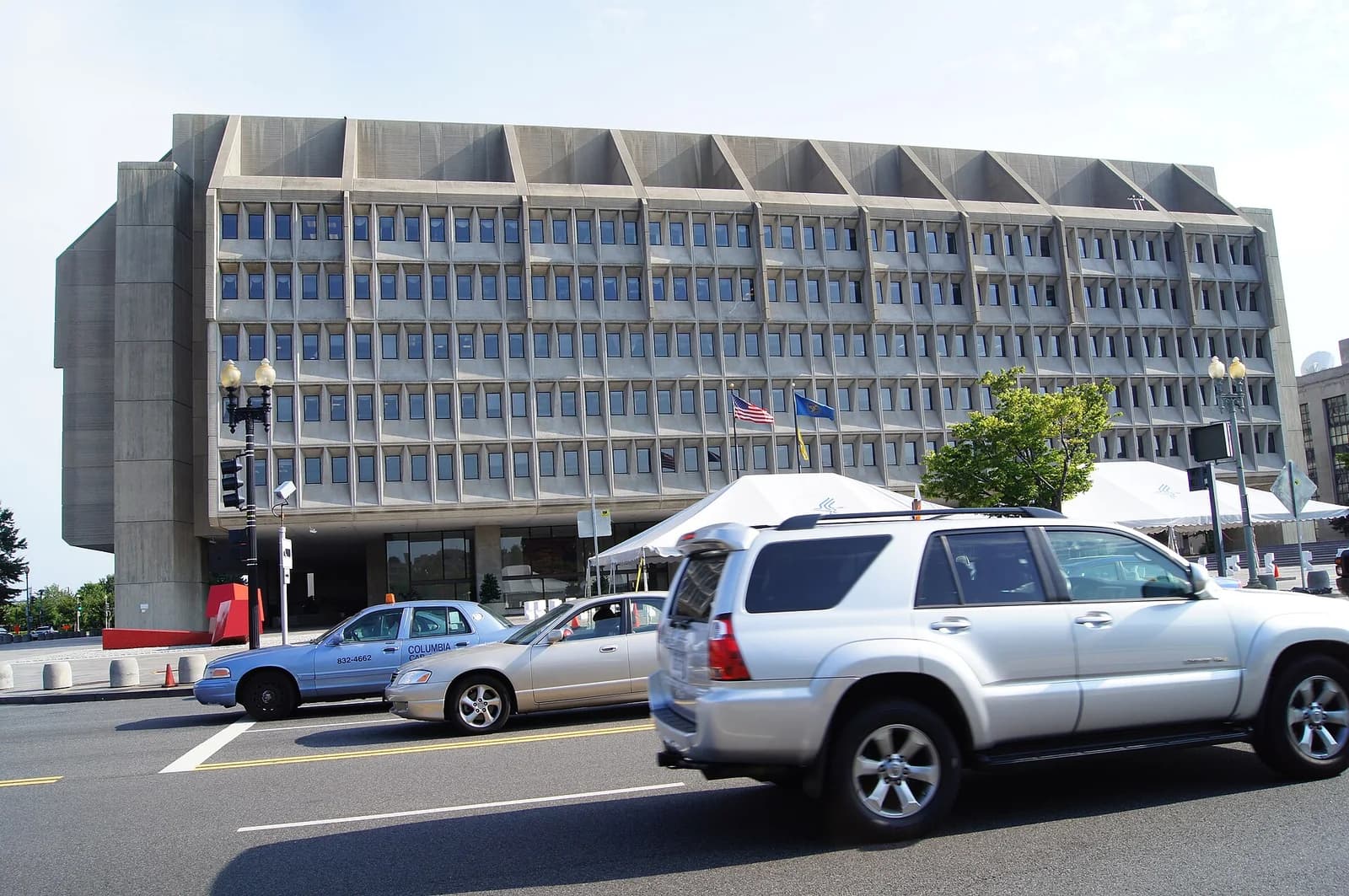 A large, brutalist-style concrete building with many small, recessed windows is seen from across a street with cars. White tents are set up in front of the building, and flags are visible near the entrance.