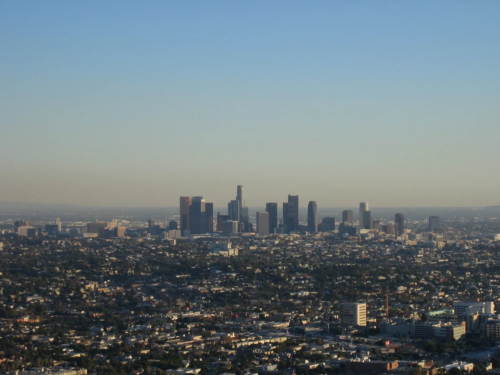 An aerial view shows a distant city skyline with numerous tall buildings rising above a sprawling urban landscape of lower residential and commercial structures. The sky is clear and light blue, with a slight haze near the horizon.