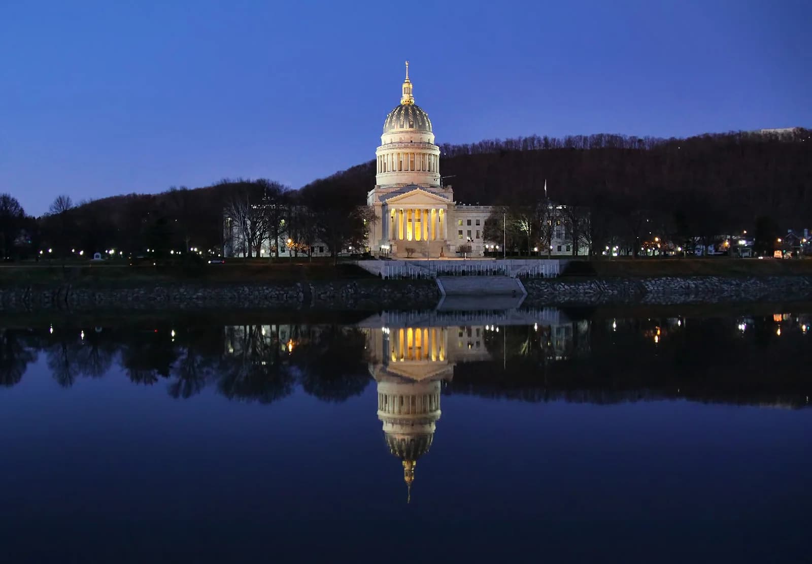 A grand, illuminated building with a large dome and columns stands on a riverbank at dusk, with its reflection visible in the dark water. A tree-covered hill rises behind the building under a deep blue sky.