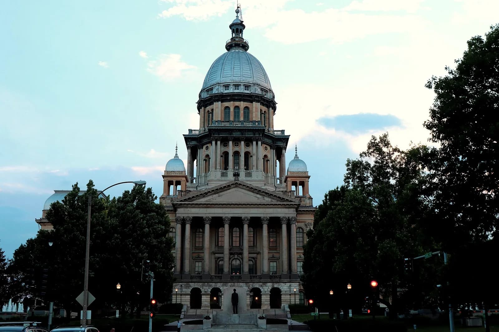 A grand, domed building with classical architecture, including columns and a large central dome, stands against a light blue sky. Dark green trees frame the lower parts of the building, and a statue is visible in front.