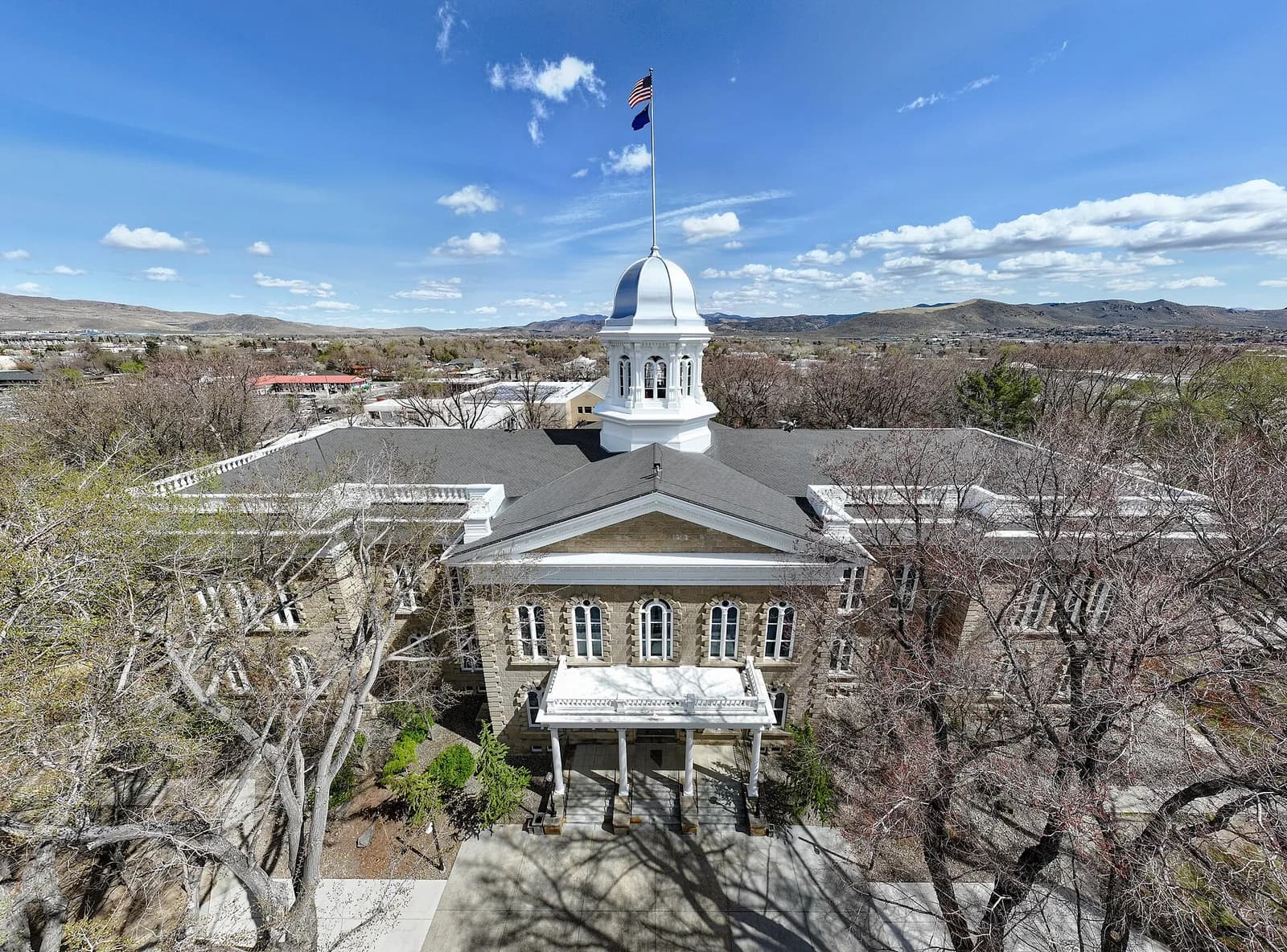 An aerial view shows a large, light-colored stone building with a dark roof and a white cupola topped with an American flag. The building is surrounded by trees, some with bare branches, under a bright blue sky with scattered white clouds.