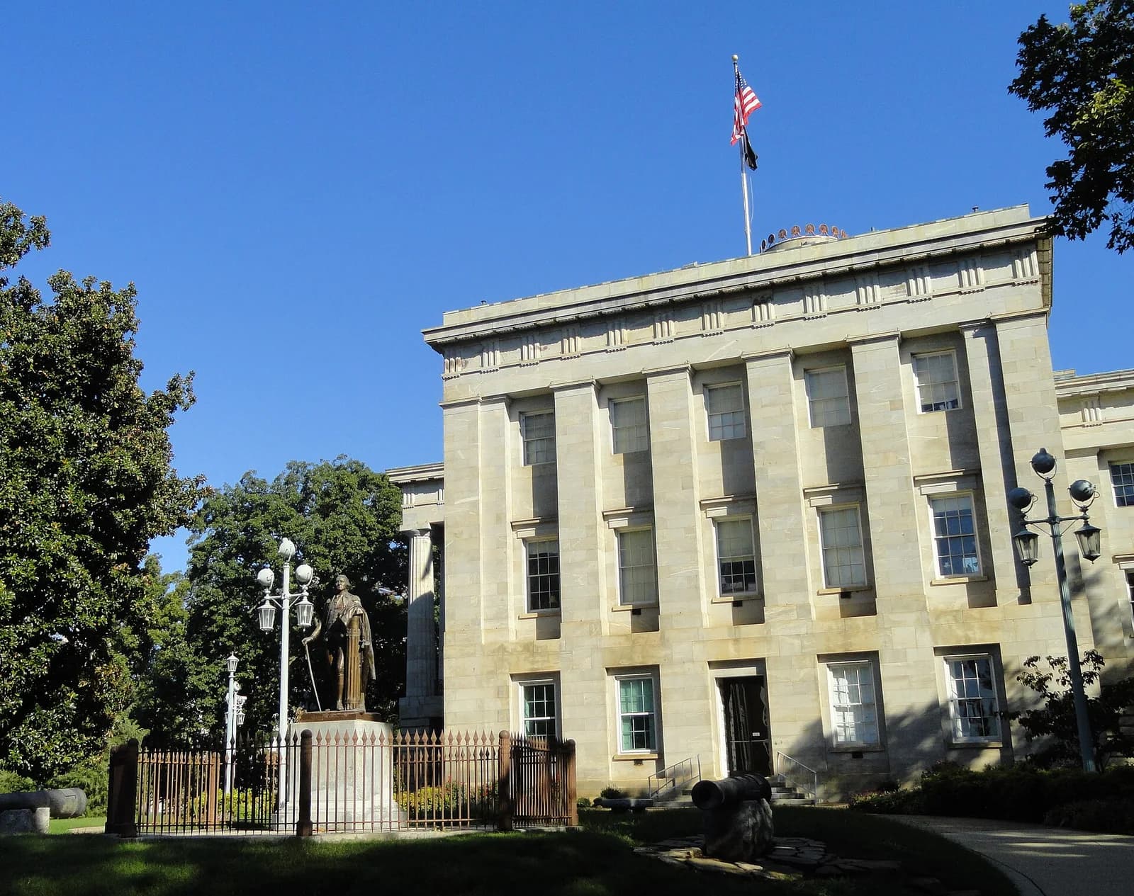 A light-colored stone building with a flat roof and an American flag flying from a flagpole on top is visible under a clear blue sky. In the foreground, a bronze statue of a man in historical attire stands behind a metal fence, surrounded by green trees and grass.