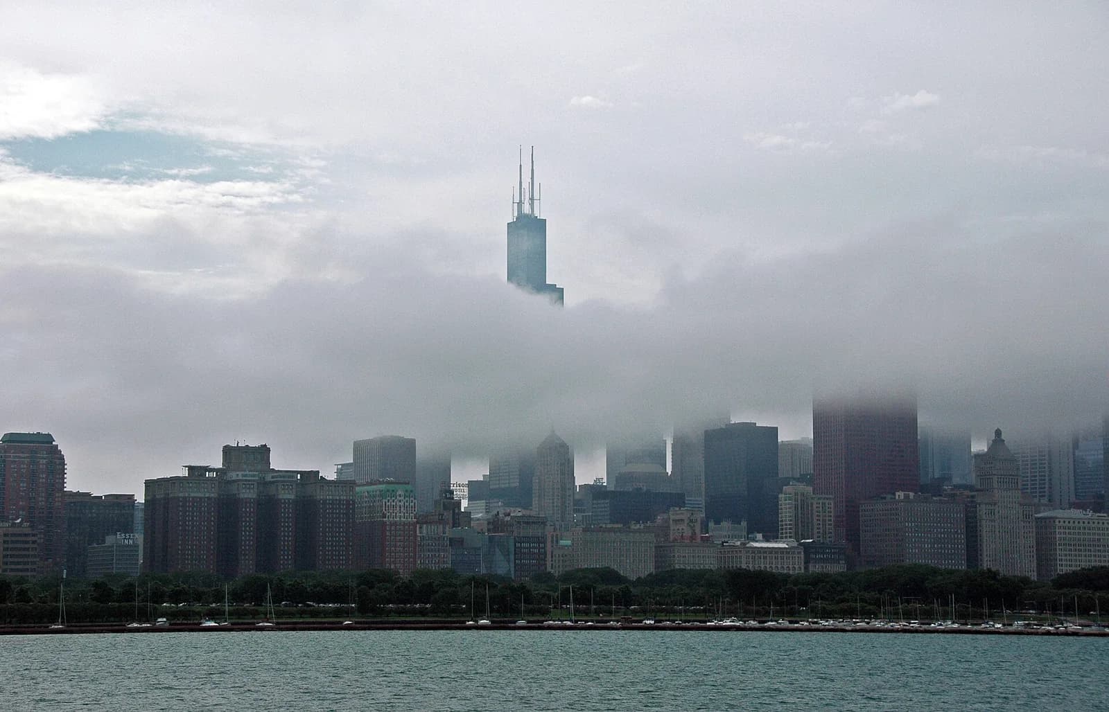 A cityscape is partially obscured by a thick layer of clouds and fog, with the top of a tall skyscraper visible above the clouds. The foreground shows a body of water with several small boats docked along the shore, and a line of trees separates the water from the buildings.