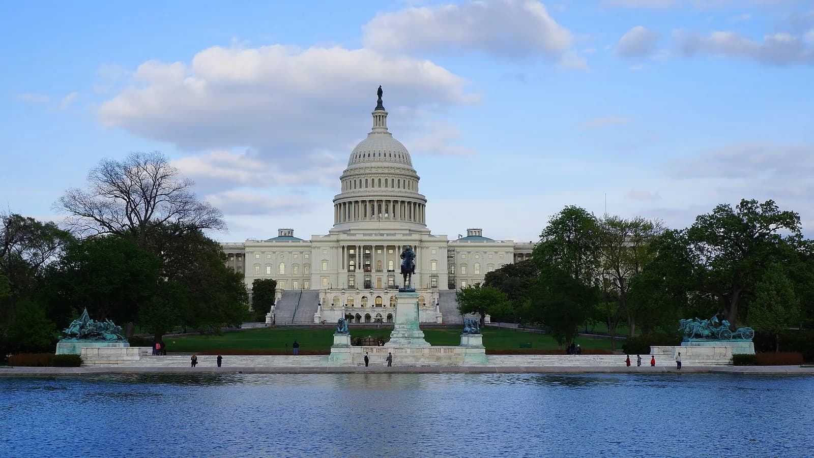 A large white domed building with columns and steps is visible across a body of water. Green trees flank the building, and several statues are visible in the foreground near the water's edge.