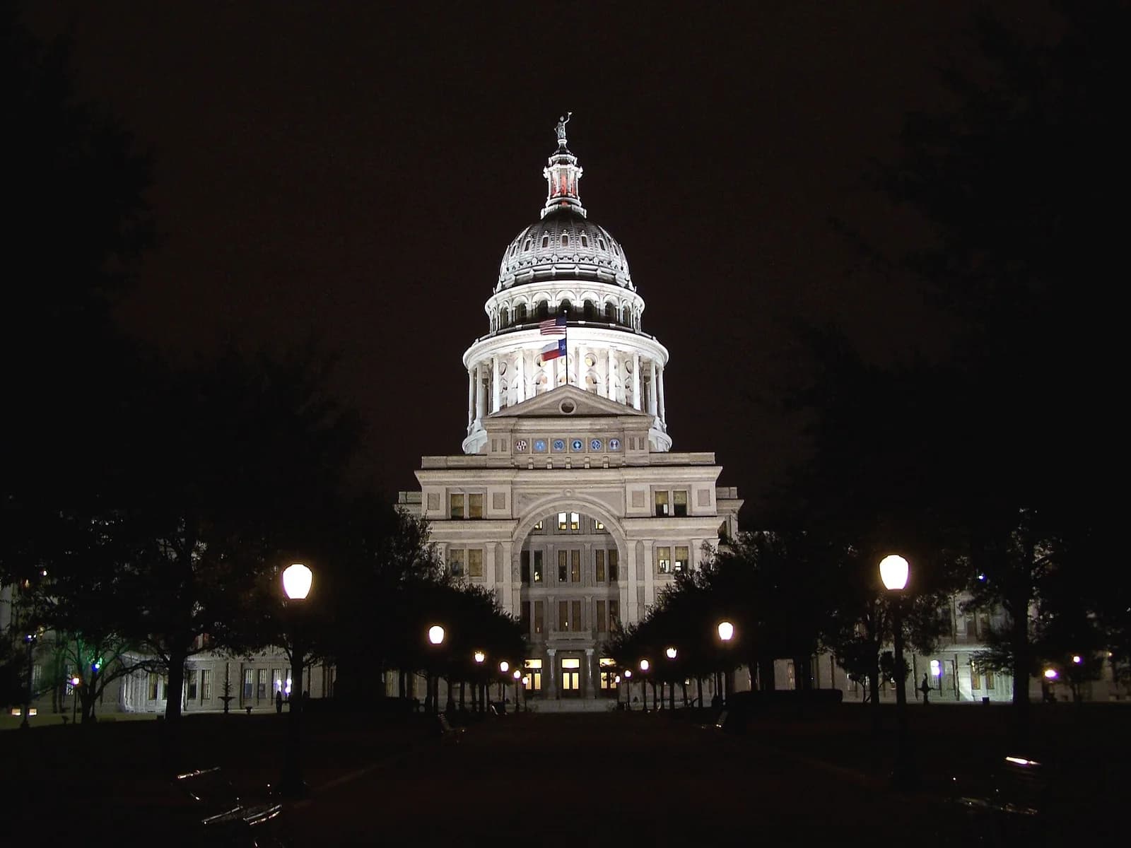 A grand, illuminated domed building stands prominently at night, with a flag visible on its facade. A tree-lined path with glowing lampposts leads up to the building, which is surrounded by dark trees and a dark sky.