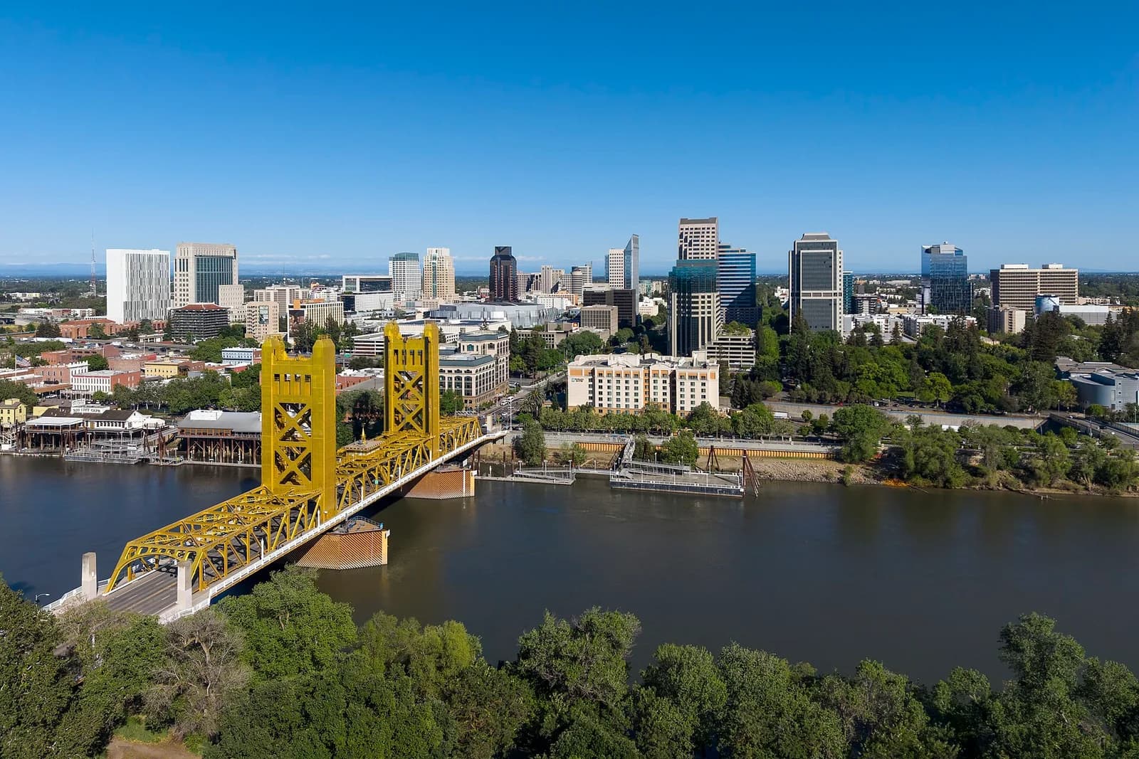 An aerial view shows a bright yellow bridge spanning a wide river, with a city skyline featuring numerous buildings and green trees on both banks under a clear blue sky.