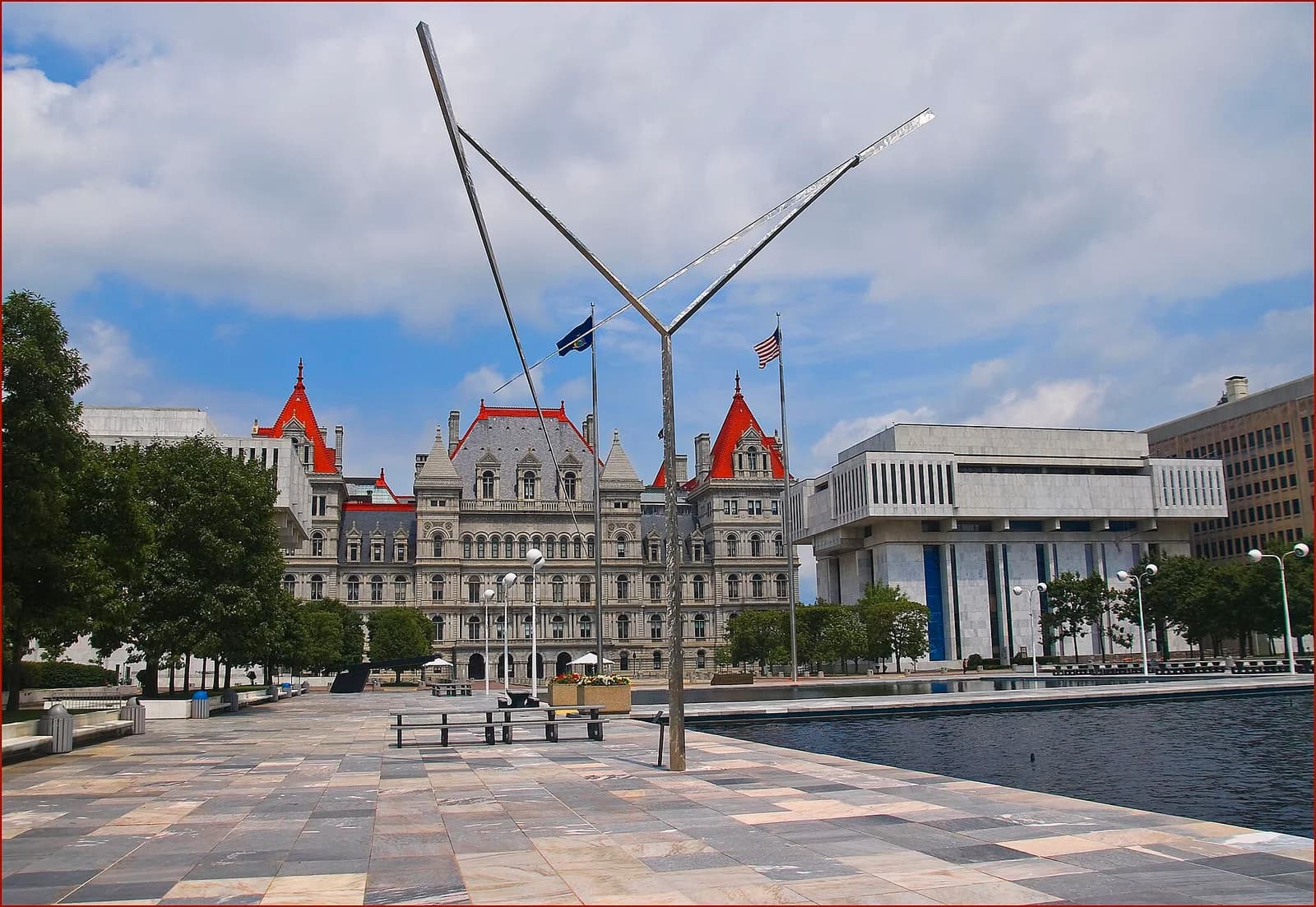 A large, ornate grey stone building with red roofs and turrets stands behind a modern plaza with a reflecting pool. A contemporary metal sculpture is in the foreground, with flags flying nearby.