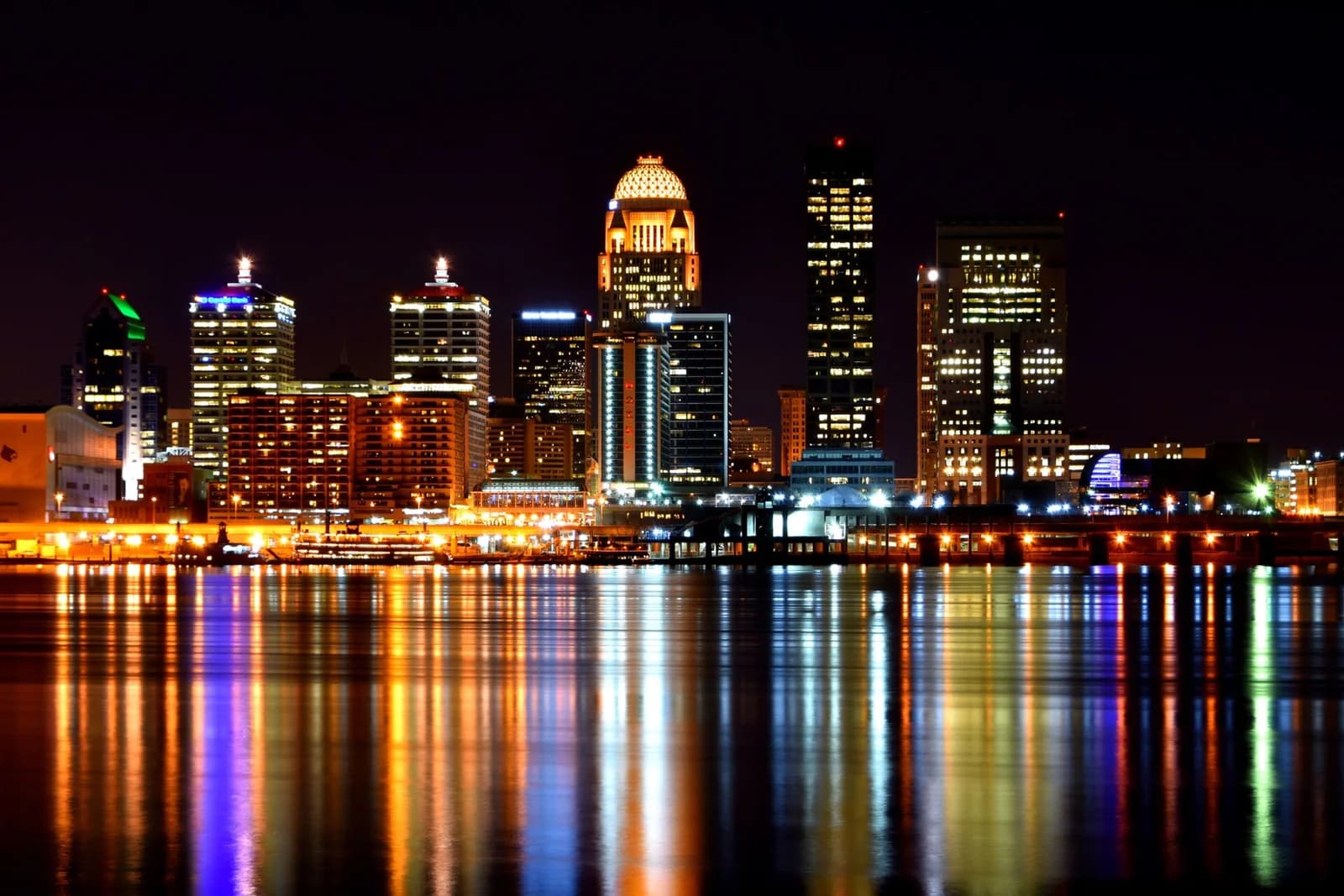 A nighttime cityscape features numerous illuminated buildings of varying heights, with a prominent domed building near the center. The city lights reflect vibrantly on the dark water in the foreground.