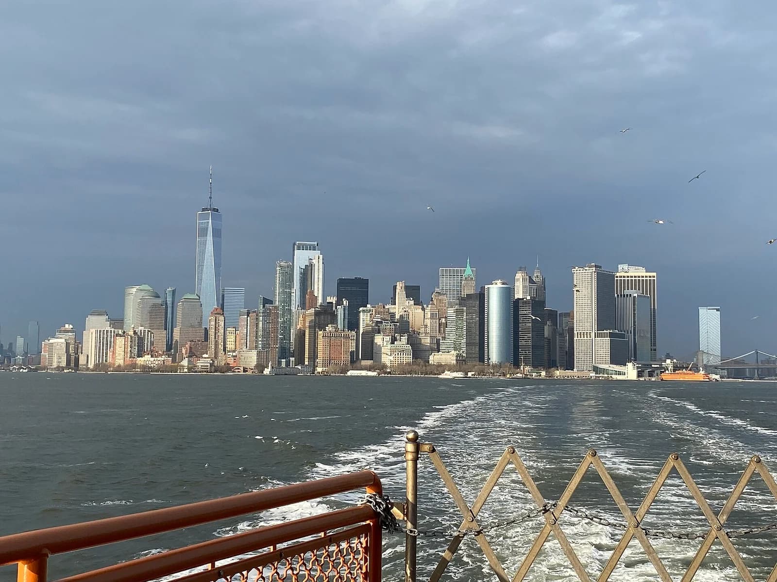 A wide view of a city skyline with numerous tall buildings under a cloudy sky, seen from the wake of a boat on a body of water. The foreground shows parts of the boat's railing and a metal gate.