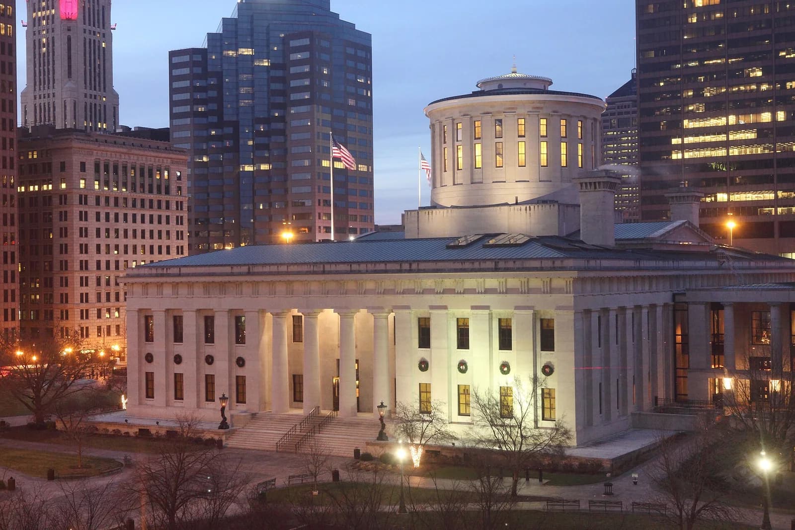An illuminated neoclassical building with a central dome and columns stands prominently in the foreground, surrounded by bare trees and streetlights. Taller, modern buildings with lit windows rise in the background under a twilight sky.