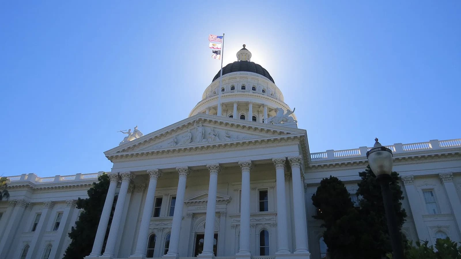 A low-angle view of a large white neoclassical building with a prominent dome and a flag flying above it. The sun is directly behind the dome, creating a bright halo effect against a clear blue sky.