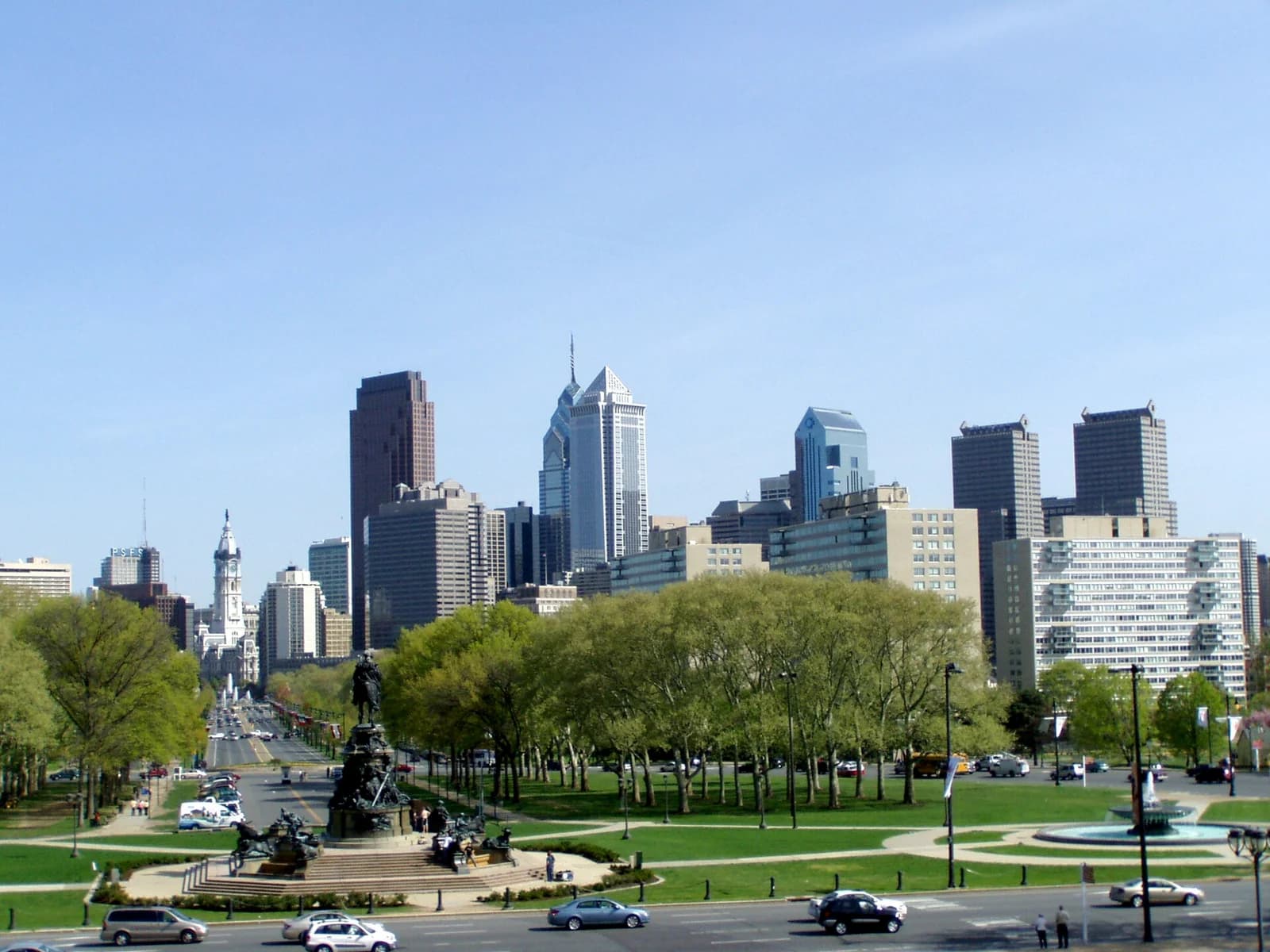 A wide view of a city skyline with numerous tall buildings under a clear blue sky. In the foreground, a green park with trees, roads, and a large equestrian statue is visible.