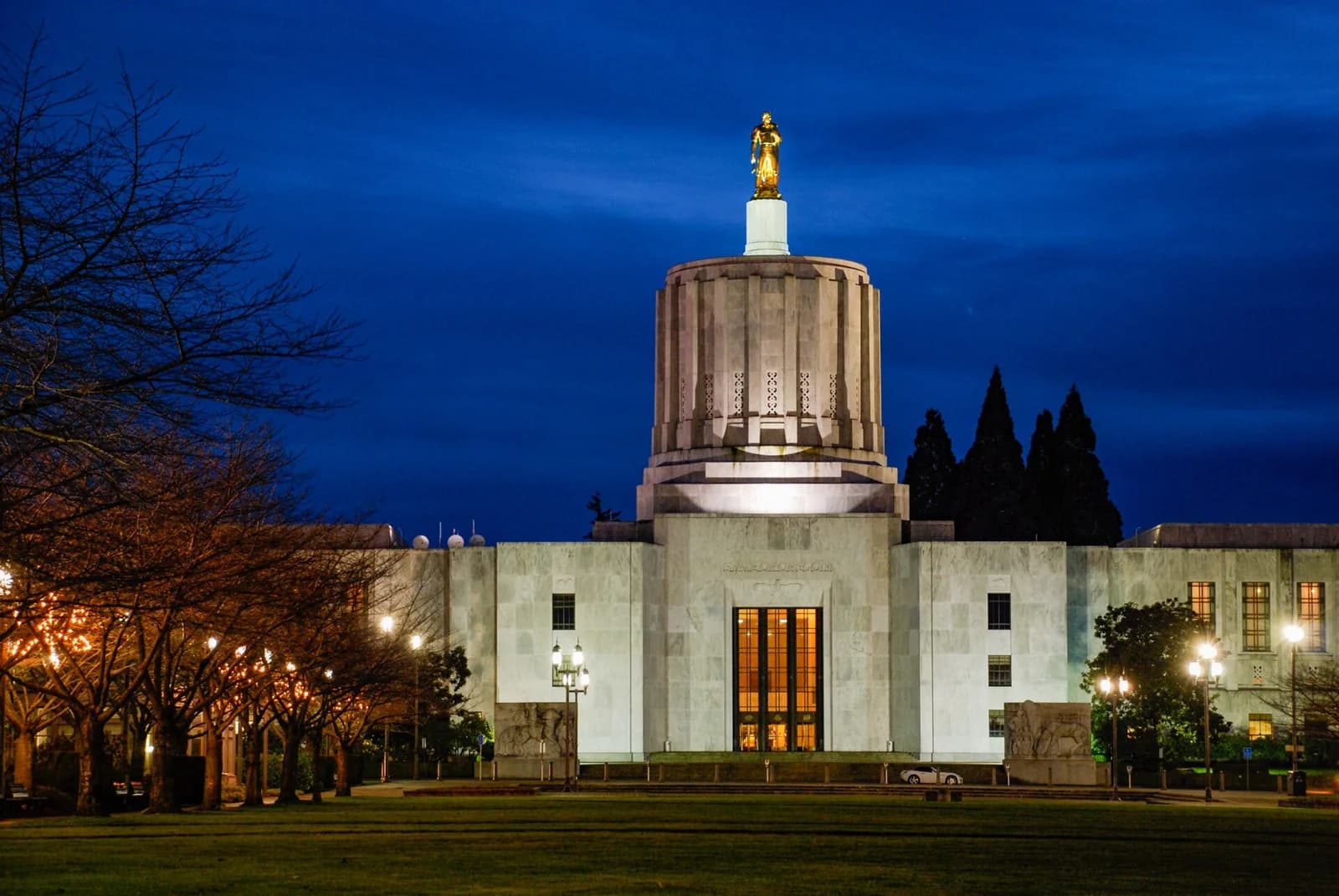 A large, light-colored institutional building with a prominent dome and a golden statue on top is illuminated at dusk. Bare trees line the left side of the building, and a grassy lawn is in the foreground.