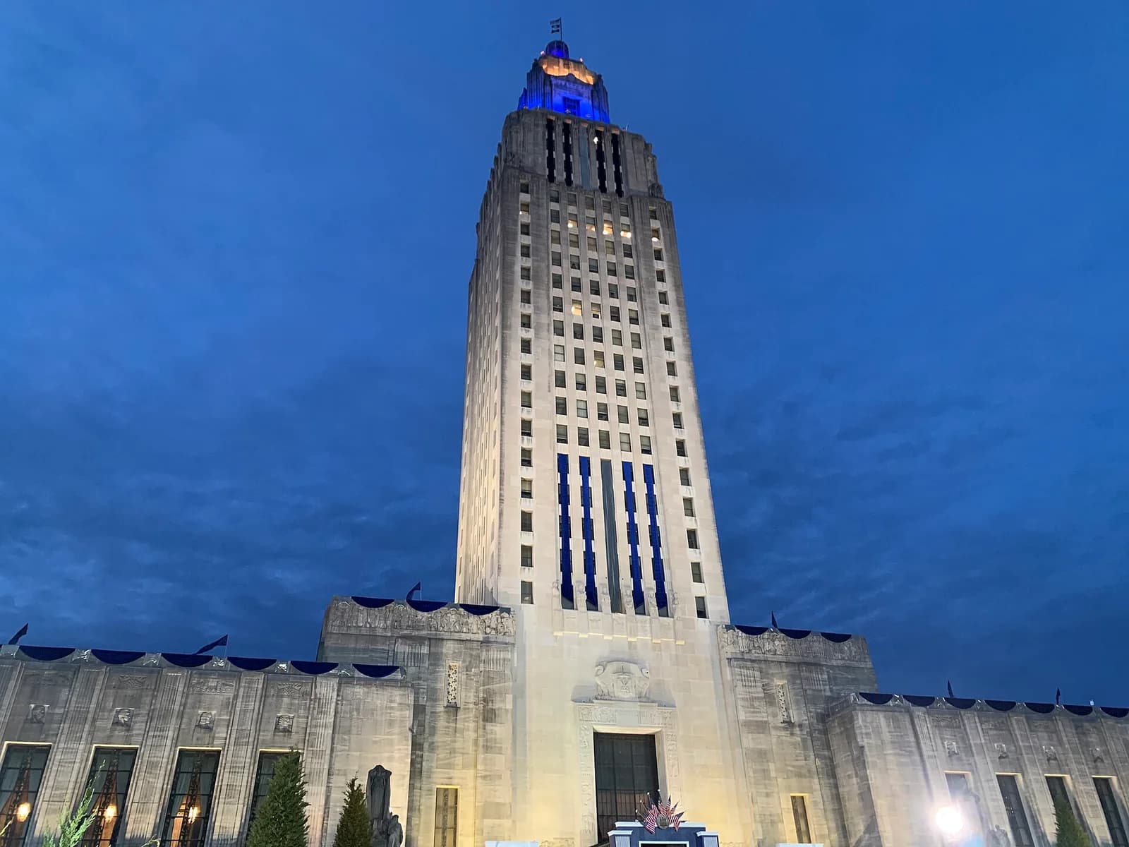 A tall, light-colored art deco building with a prominent central tower is illuminated with blue lights against a dark blue evening sky. The lower sections of the building feature decorative elements and multiple windows, with some blue banners draped along the roofline.