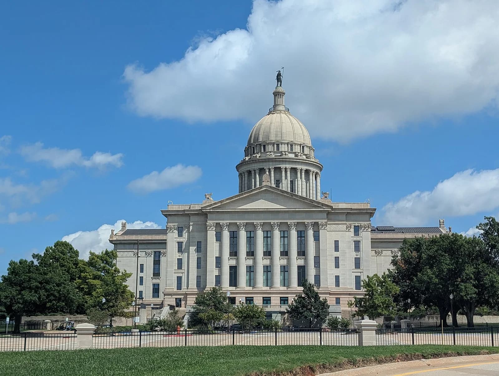 A grand, light-colored stone building with a large dome topped by a statue stands under a blue sky with scattered white clouds. The building features classical architectural elements, including columns and a prominent portico, and is surrounded by green grass and trees, with a black fence in the foreground.