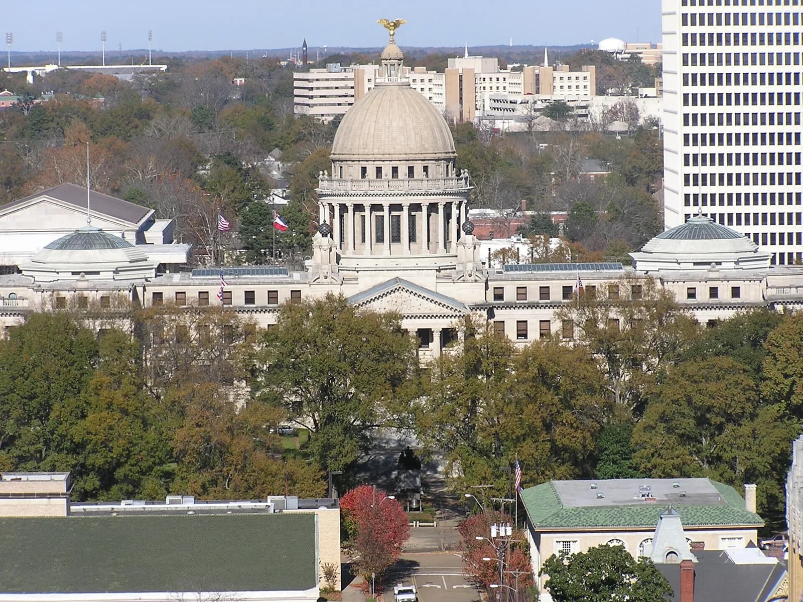 An aerial view shows a large, light-colored neoclassical building with a central dome topped by a golden eagle, flanked by two smaller domes. The building is surrounded by green trees, with other urban buildings visible in the background under a clear sky.