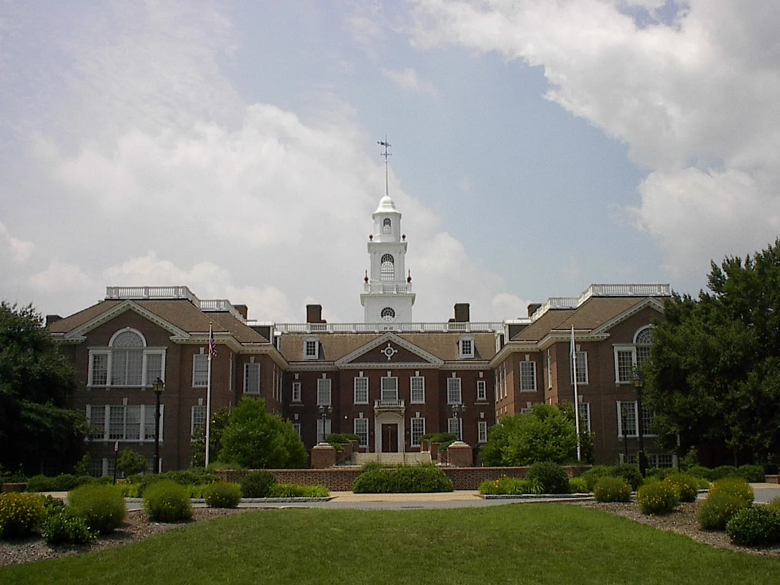 A large, symmetrical brick building with a white cupola and spire stands under a partly cloudy sky. The building is surrounded by green lawns and manicured bushes.