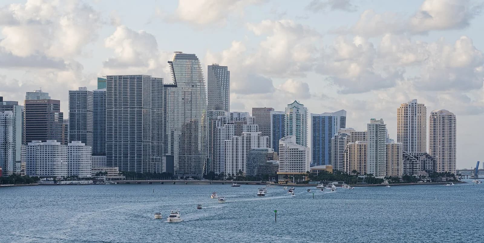 A wide shot of a city skyline with numerous tall buildings and skyscrapers, some with distinctive architectural features, under a cloudy sky. In the foreground, a body of water is dotted with several small boats, leaving wakes behind them.