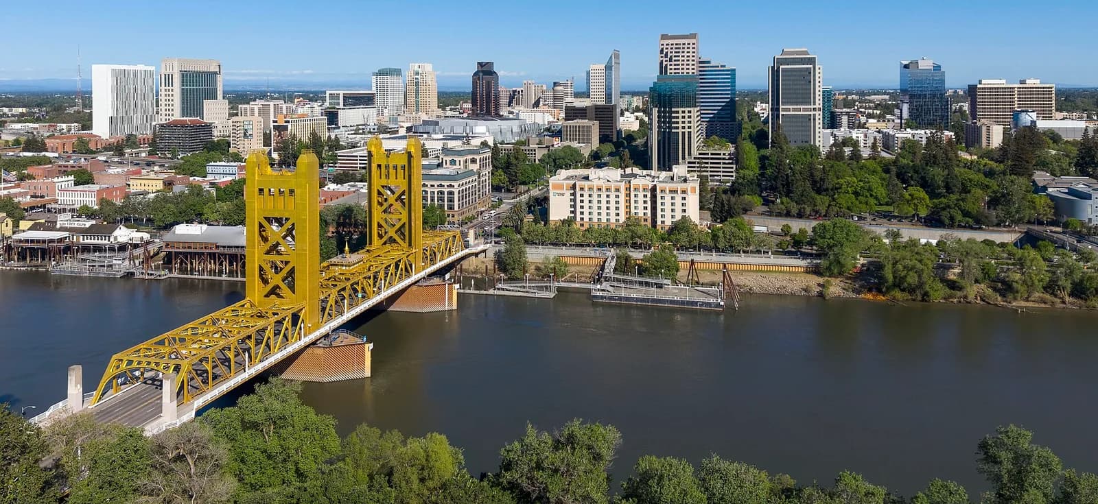 An aerial view shows a bright yellow bridge spanning a wide river, with a city skyline featuring numerous tall buildings in the background under a clear blue sky. Lush green trees line both sides of the river and are interspersed among the buildings.