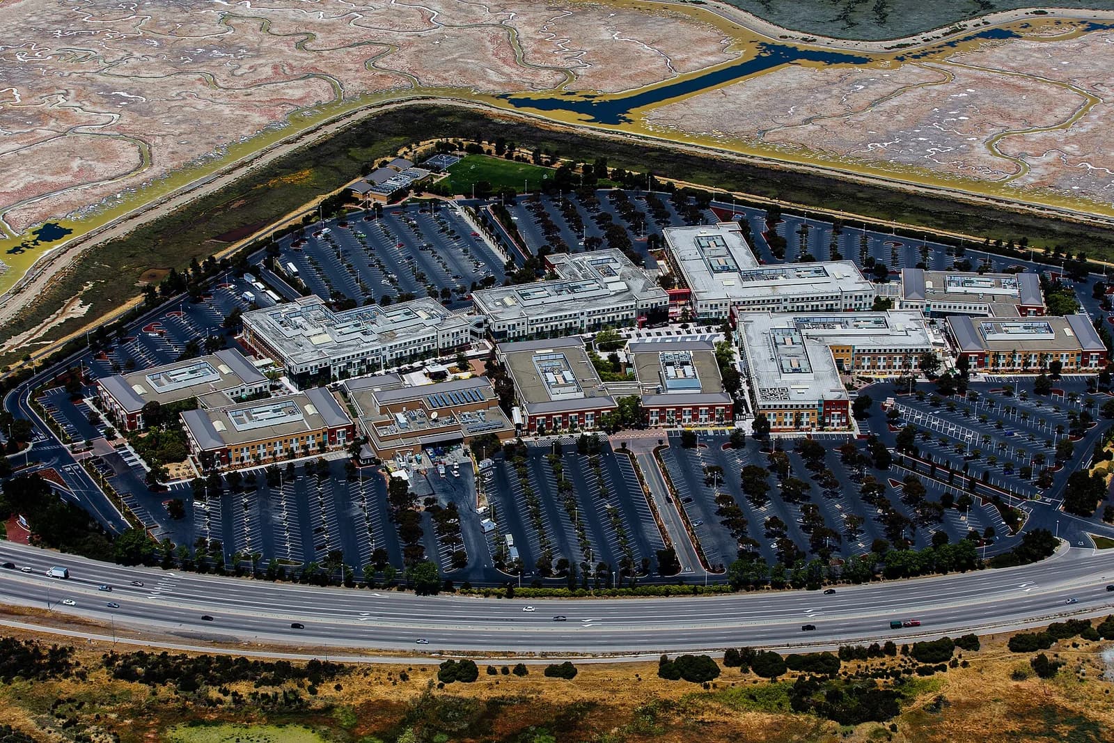 An aerial view shows a large complex of buildings with extensive parking lots, surrounded by a highway and natural wetlands. The buildings feature a mix of light-colored and orange-brown facades with numerous windows.
