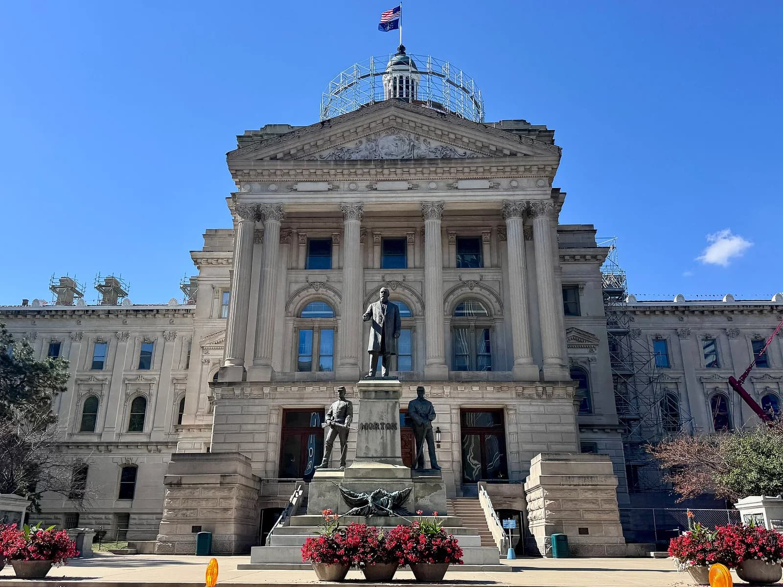 A large, ornate stone building with a central dome and columns stands under a clear blue sky. In front of the building, a monument with several statues is visible, flanked by large planters with red flowers.