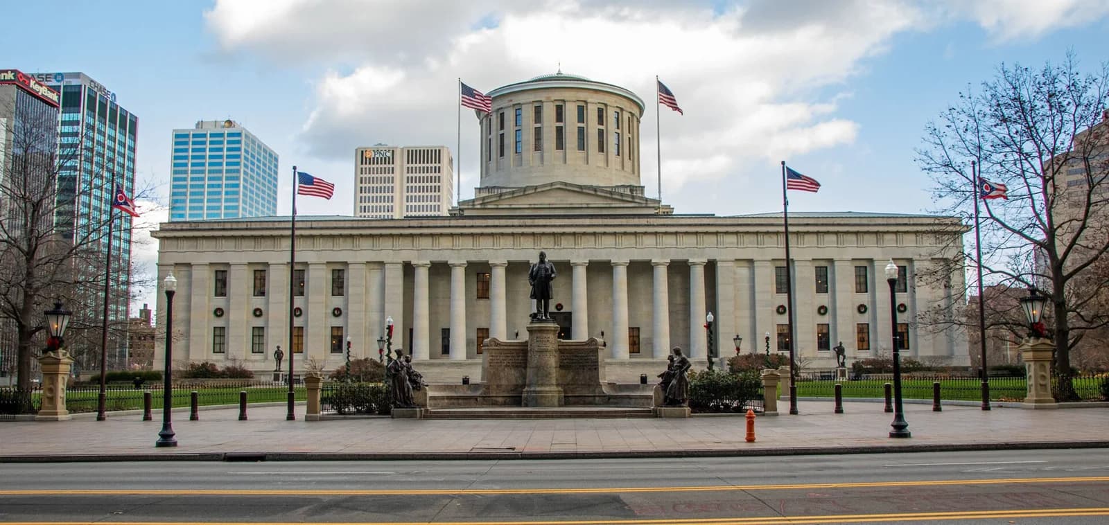 A large, light-colored stone building with a central dome and columns stands behind a statue and a grassy lawn. American flags fly on poles in front of the building, with other tall buildings visible in the background.