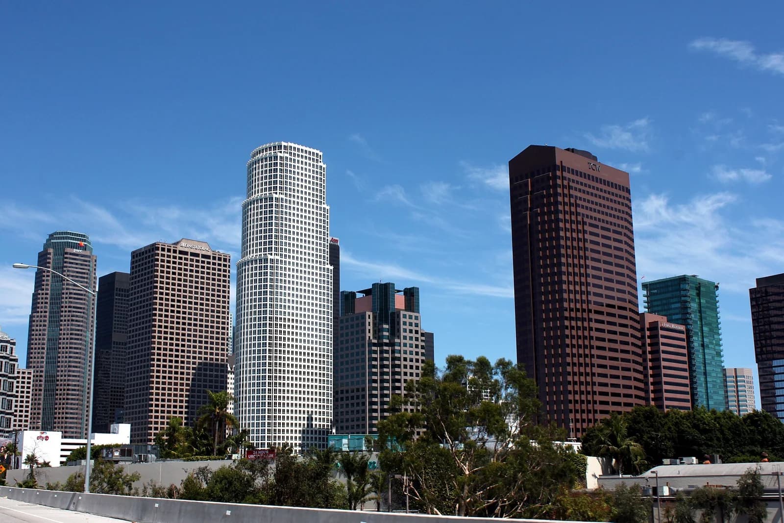 A cityscape features several tall buildings under a clear blue sky with wispy clouds. The foreground shows a concrete barrier and green foliage, suggesting a view from a highway or elevated road.