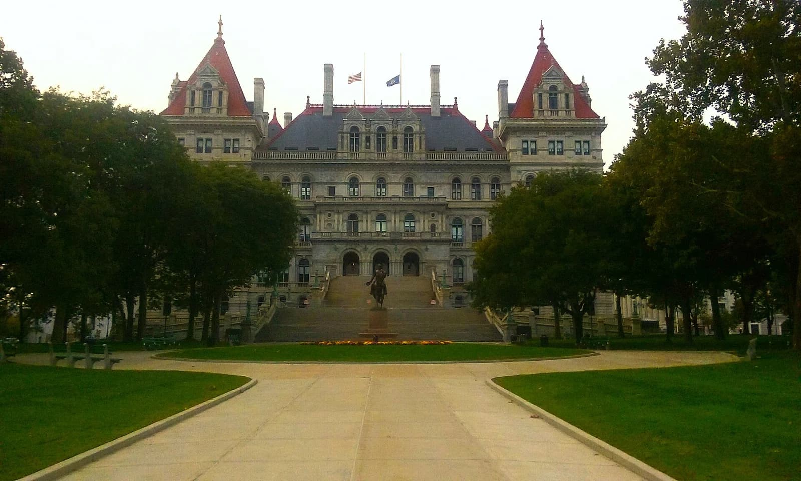 A grand, ornate stone building with a dark roof and red-capped turrets stands behind a wide, green lawn. A statue of a person on horseback is visible at the base of the building's steps.