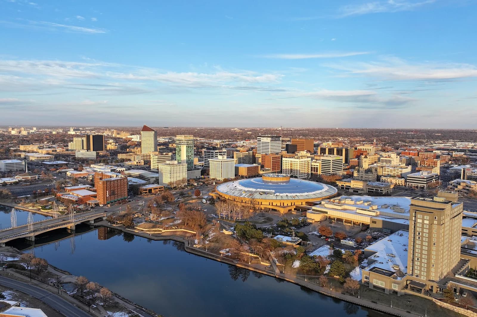 An aerial view of a city shows a river winding through the urban landscape, with numerous buildings and a large domed structure. Some snow is visible on the ground and rooftops, under a clear blue sky with wispy clouds.