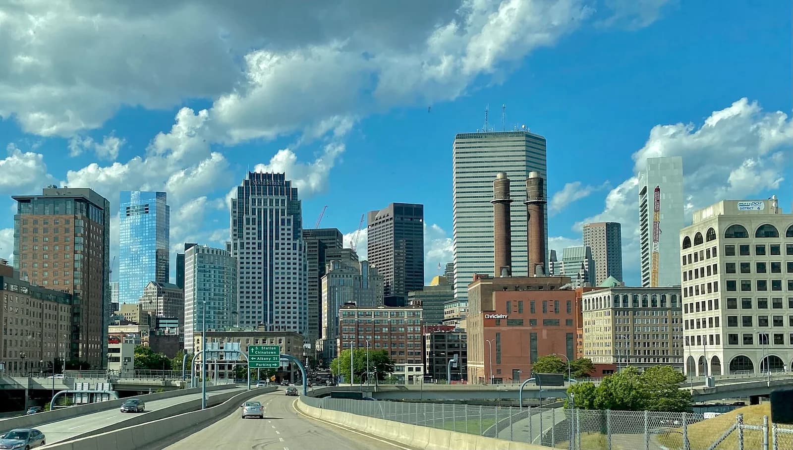 A wide view of a city skyline with numerous tall buildings under a blue sky with white clouds. In the foreground, a multi-lane highway curves, with several cars visible.
