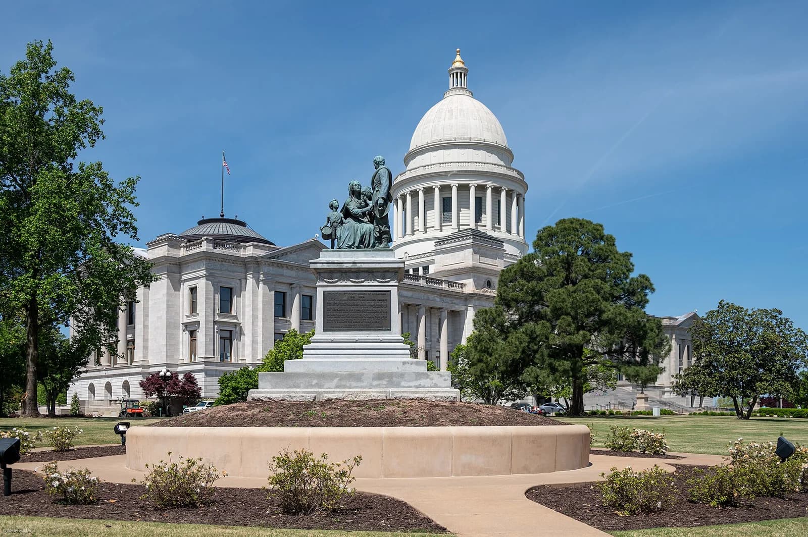 A large, light-colored domed building with columns stands under a clear blue sky. In the foreground, a bronze statue on a stone pedestal is surrounded by landscaped grounds with trees and bushes.