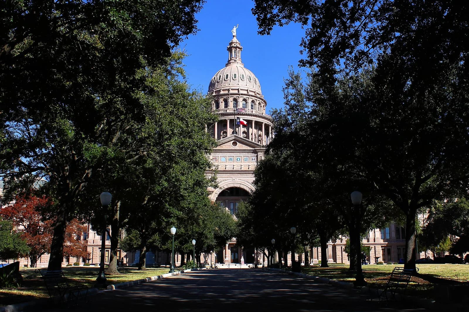A grand domed building with a statue on top is framed by dark green trees on either side of a paved path. The building is light-colored with columns and flags flying from its dome.