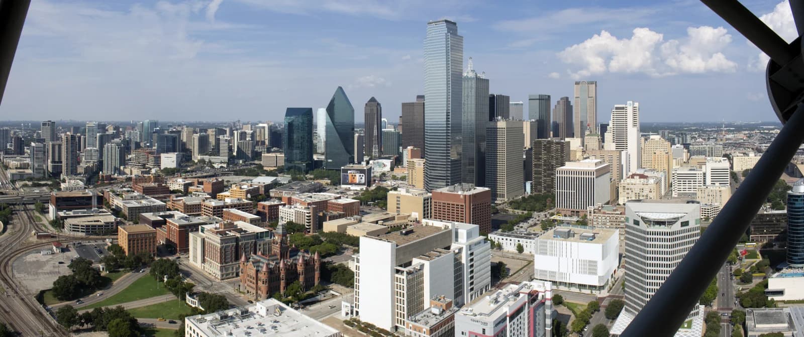 An aerial panoramic view of a city skyline under a partly cloudy sky, featuring numerous high-rise buildings and a network of roads and railway tracks. A dark structural element is visible in the foreground on the right.
