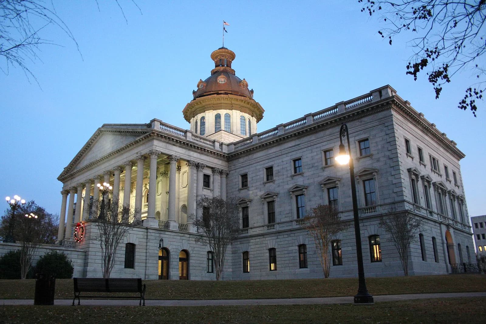 A large, classical-style building with a prominent dome and columns is shown at dusk. The building is made of light-colored stone and is surrounded by a grassy lawn with a few trees and a streetlamp.
