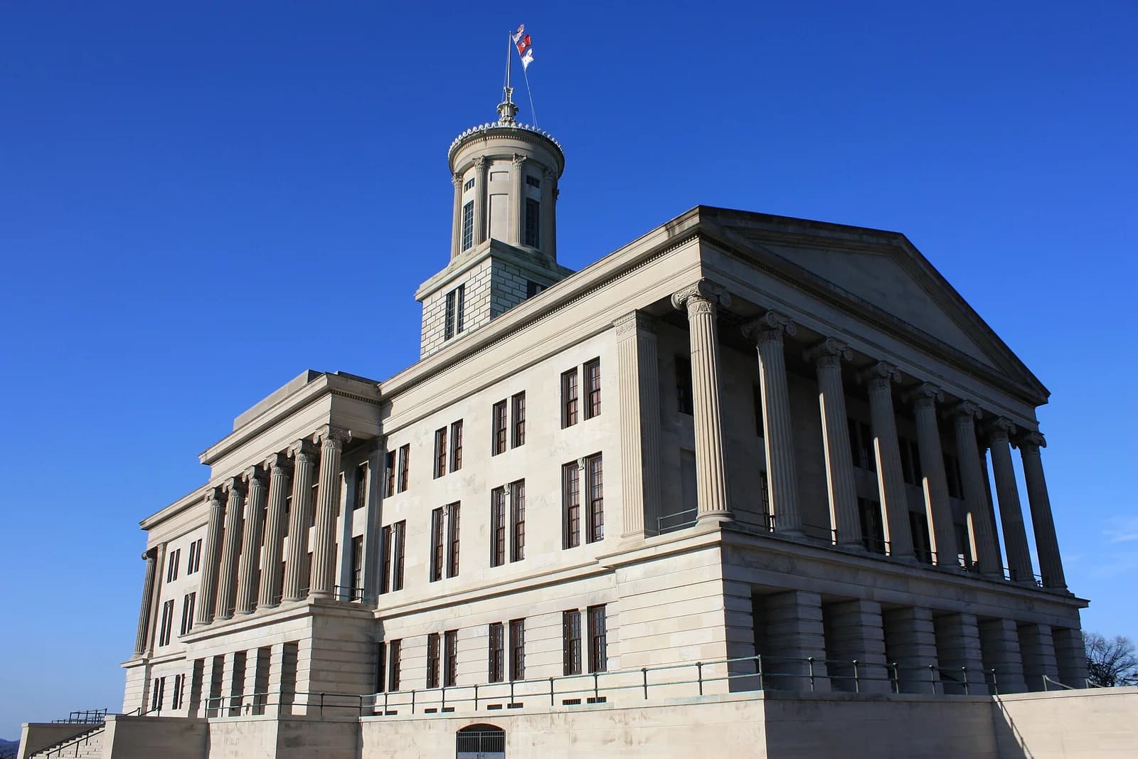 A large, light-colored stone building with classical architectural elements, including columns and a central cupola with a flag, stands against a clear blue sky. The building is viewed from a low angle, showing its full facade and part of its base.