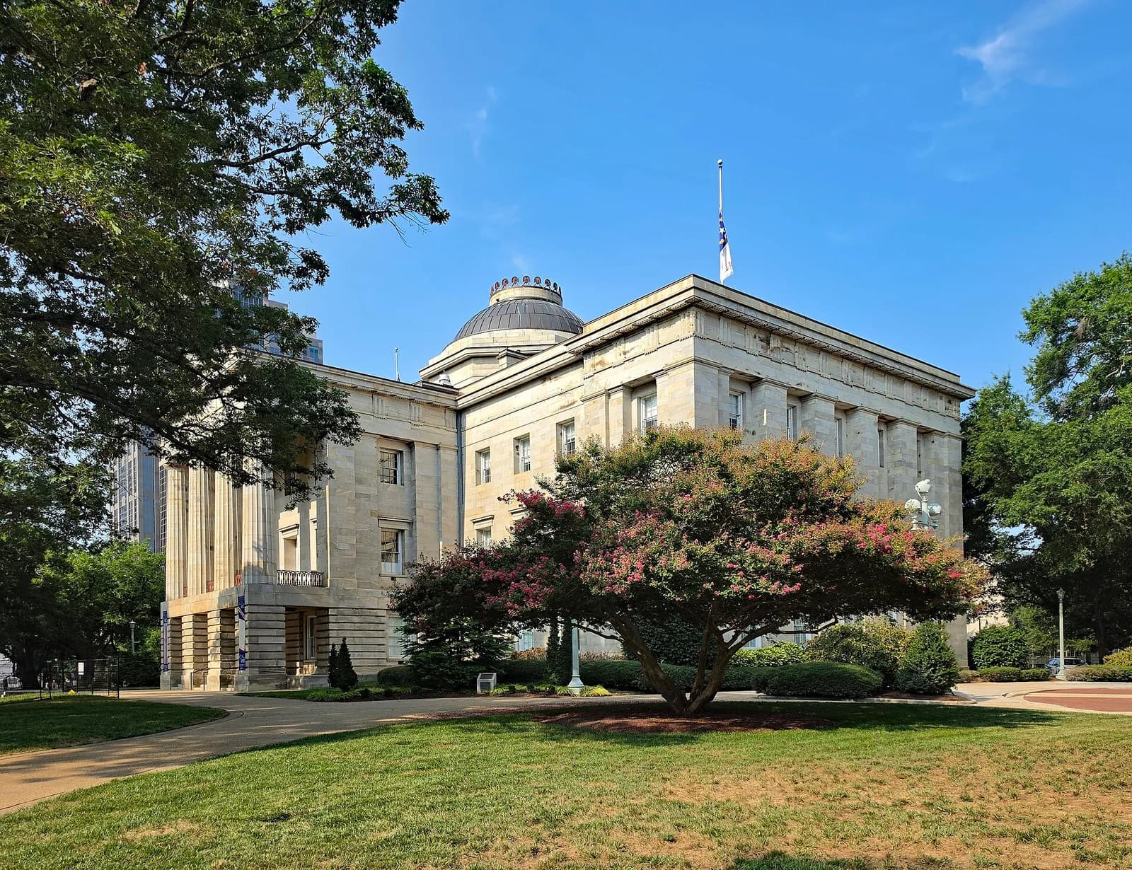 A large, light-colored stone building with a dome and columns stands under a clear blue sky. Green trees and a flowering bush are in the foreground, with a paved path and grass leading up to the building.