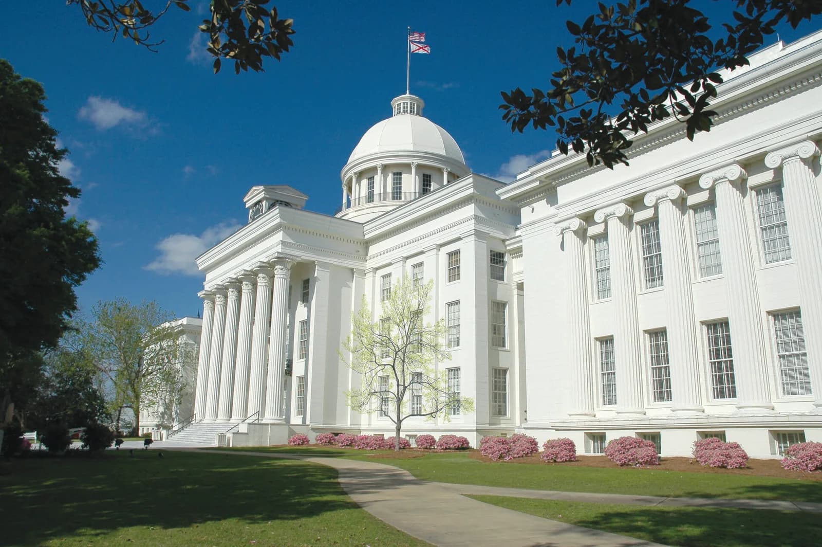 A large white building with a dome and columns stands under a clear blue sky. A paved path curves through a green lawn with pink flowering bushes in the foreground.