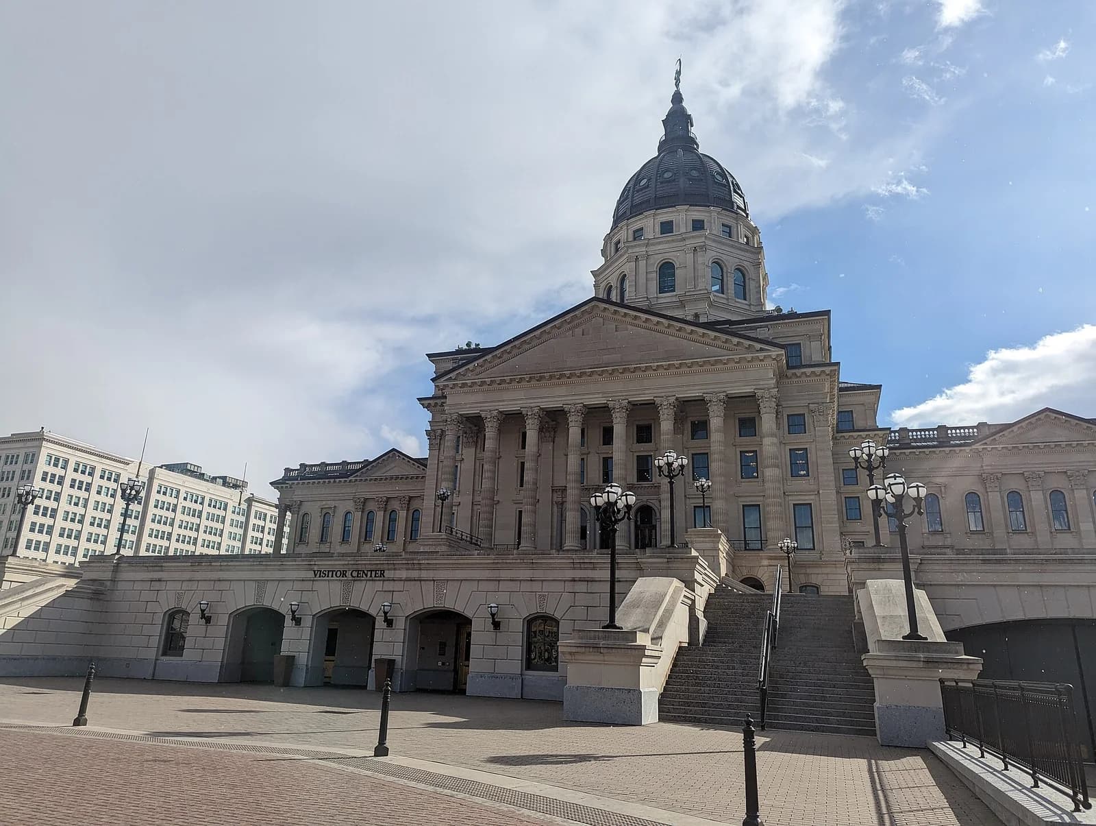 A large, light-colored stone building with a prominent dome and columns stands under a partly cloudy sky. A wide staircase leads up to the main entrance, and a "VISITOR CENTER" sign is visible on the lower level.