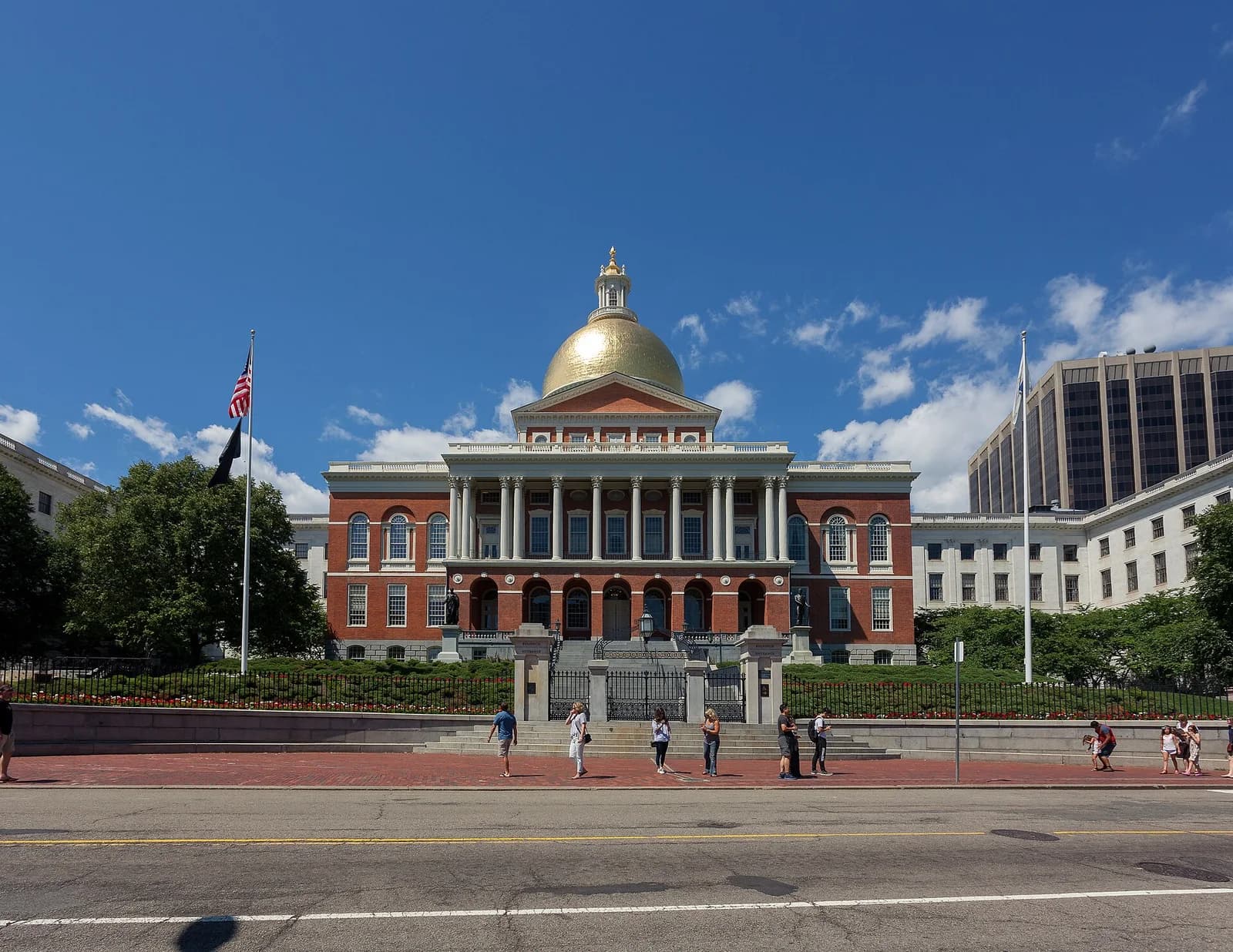 A large red brick building with a golden dome and white columns stands under a clear blue sky with scattered clouds. People are walking on the sidewalk in front of a black fence and landscaped area, with a street in the foreground.
