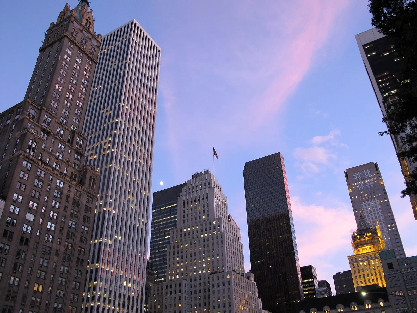 A low-angle view of a cityscape at dusk shows several tall buildings against a sky with pink and blue hues. A crescent moon is visible between two of the buildings.