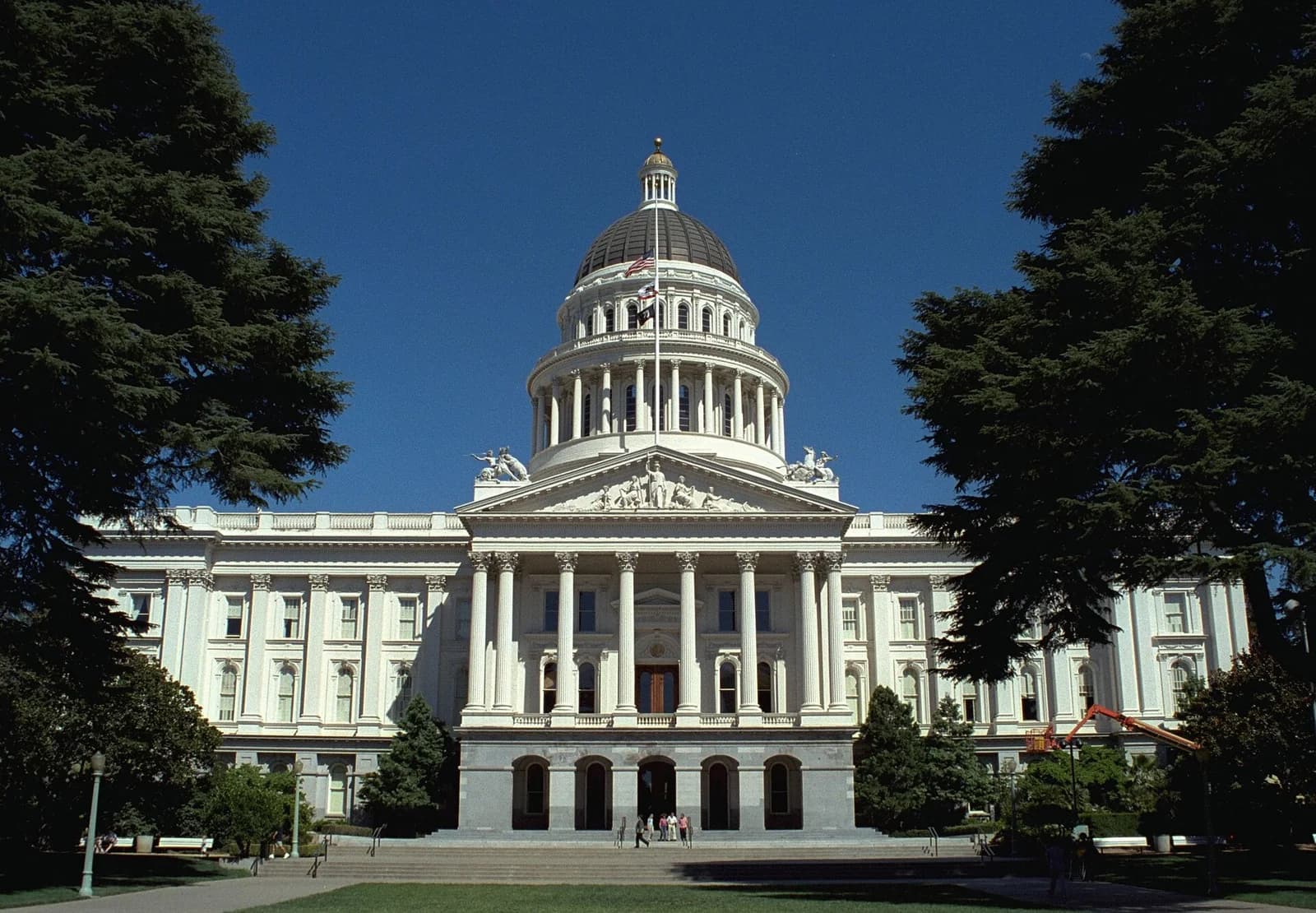 A grand white neoclassical building with a large dome and columns stands prominently against a clear blue sky. Lush green trees frame the building on both sides, with a manicured lawn and steps leading up to the entrance.