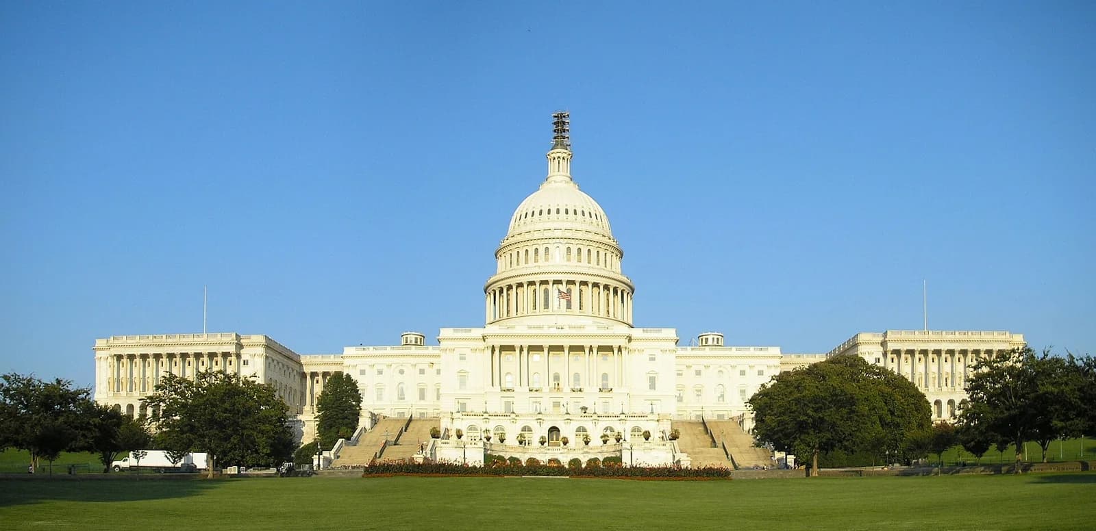A large, white neoclassical building with a prominent dome and columns stands under a clear blue sky. Green lawns and trees are visible in the foreground and on either side of the building.