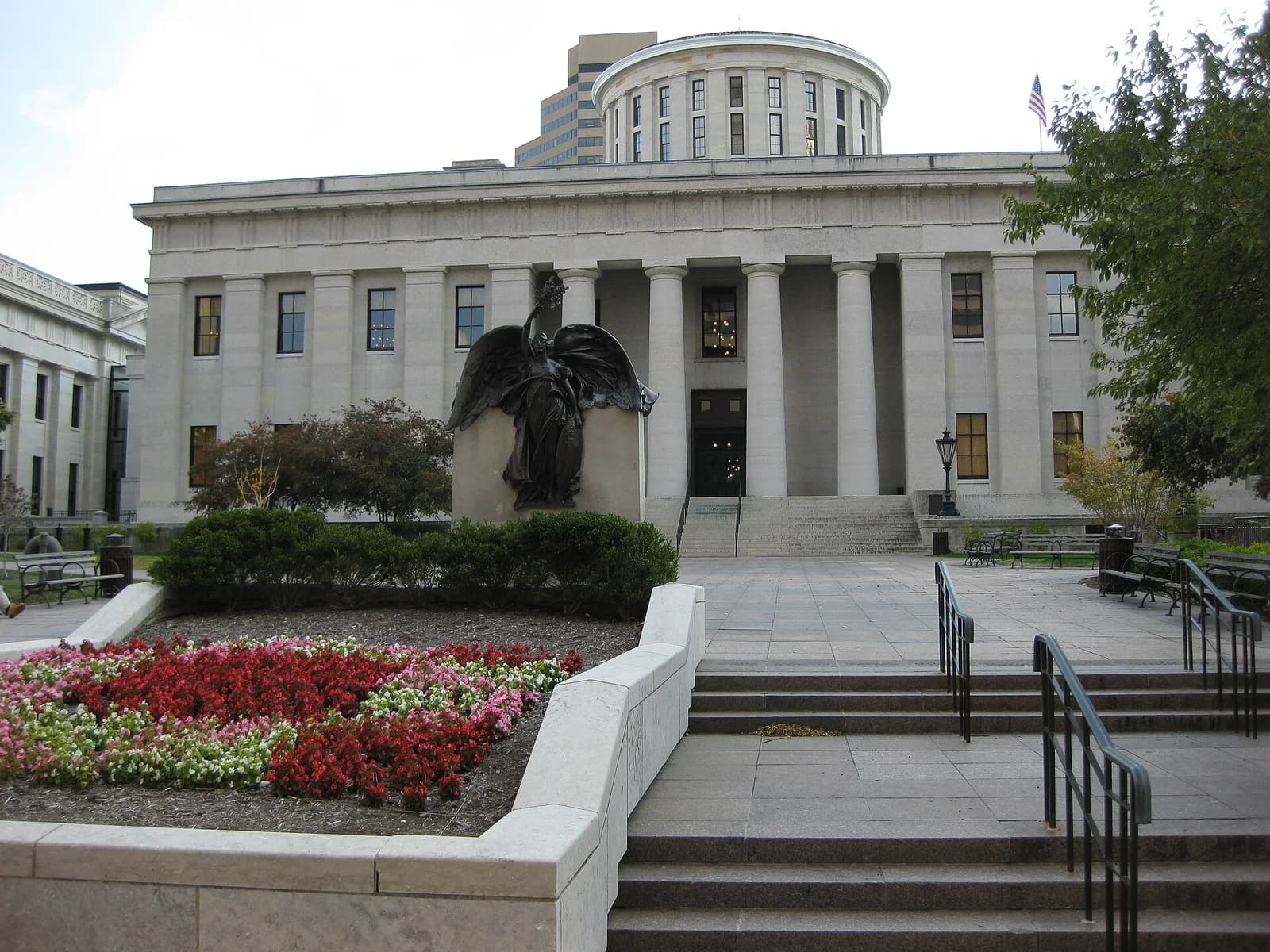 A large, light-colored stone building with a domed roof and columns stands behind a plaza with steps, flowerbeds, and a dark statue. An American flag flies from the building's roof.