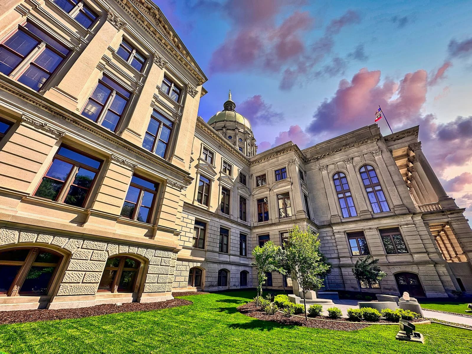 A grand, light-colored stone building with a golden dome and a flag flying from its roof is seen from a low angle. The building features many windows, columns, and ornate architectural details, with green grass and small trees in the foreground under a dramatic sky with pink and blue clouds.